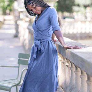 Woman in a soft blue linen wrap dress with V-neckline and tie waist, standing gracefully by a sunlit stone railing. The dress features a flattering midi length and elbow-length sleeves, with a delicate handmade decorative stitch along the neckline that highlights its artisanal quality. Ideal for summer vacations, city strolls, brunches, or garden gatherings. This elegant dress is ready to ship from the USA within one business day.