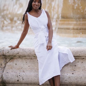 Woman in a white linen wrap dress with a wide waist tie, sitting gracefully by a historic fountain. The sleeveless V-neck dress flows naturally with a slit, offering a light, airy feel perfect for summer getaways, destination weddings, or elegant resort evenings. Crafted from breathable linen and styled with minimal accessories. Ready to ship from the USA in 1 business day.
