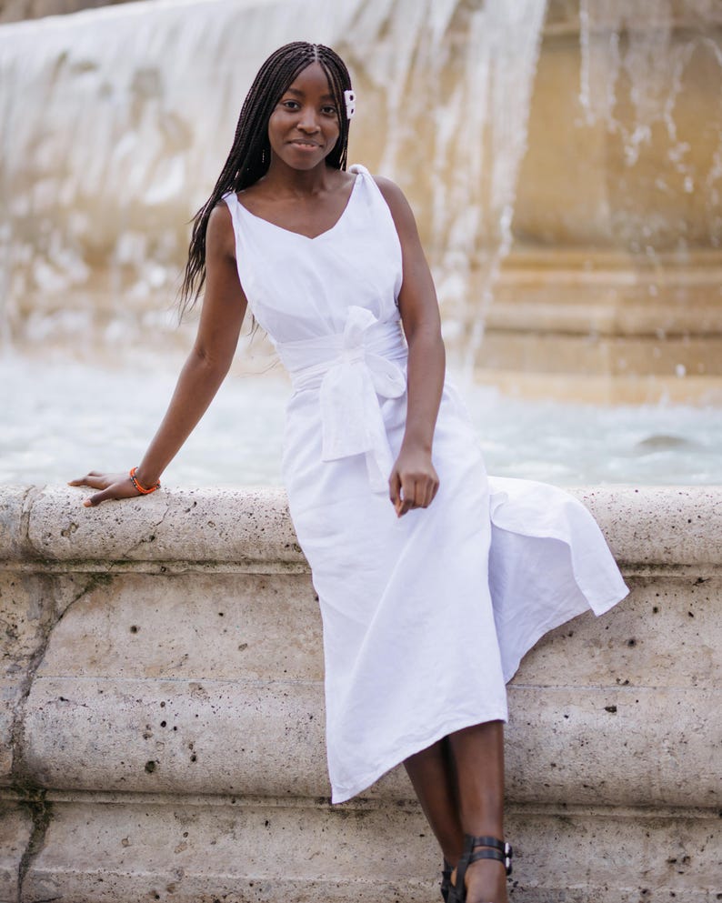 Woman in a white linen wrap dress with a wide waist tie, sitting gracefully by a historic fountain. The sleeveless V-neck dress flows naturally with a slit, offering a light, airy feel perfect for summer getaways, destination weddings, or elegant resort evenings. Crafted from breathable linen and styled with minimal accessories. Ready to ship from the USA in 1 business day.