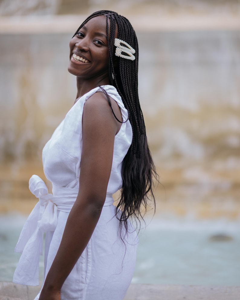 Smiling woman in a white linen wrap dress with a feminine bow-tie waist, photographed from the side. The dress features a V-back, wide straps, and a flattering silhouette made of breathable natural linen. Perfect for summer vacations, beach dinners, honeymoons, or minimalist weddings. A joyful, romantic look ideal for confident women who value comfort and elegance. Ships fast from the USA within 1 business day.