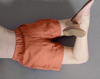 May include: Rust-colored linen shorts with an elastic waistband. The shorts are paired with a light-colored top and ballet flats. The flats are a soft pink color. The image is shot against a gray background.