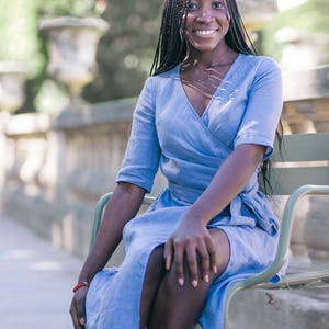 Smiling woman seated on a green chair, wearing a light blue linen wrap dress with a V-neck and tie waist. The dress features a feminine silhouette with elbow-length sleeves and a soft midi hem, accented by a handmade decorative stitch along the neckline. Ideal for summer travel, city vacations, garden lunches, or casual workdays. Ships fast from the USA within one business day.