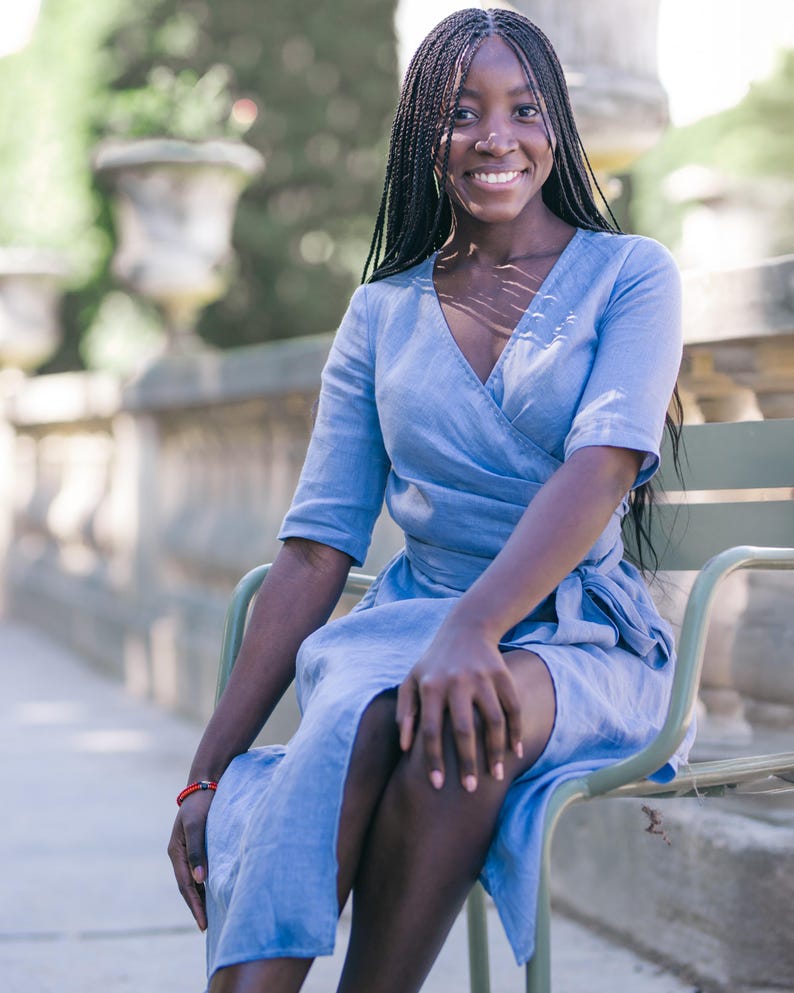Smiling woman seated on a green chair, wearing a light blue linen wrap dress with a V-neck and tie waist. The dress features a feminine silhouette with elbow-length sleeves and a soft midi hem, accented by a handmade decorative stitch along the neckline. Ideal for summer travel, city vacations, garden lunches, or casual workdays. Ships fast from the USA within one business day.