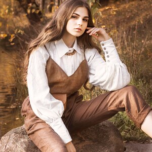 Woman wearing a white romantic linen blouse with puff sleeves, button cuffs, and vintage collar, styled with brown overalls in a fall forest setting. Victorian-inspired button-up shirt perfect for cottagecore outfits, feminine vintage looks, or countryside style. Ideal for photoshoots, layering, or holiday fashion. Ships fast from Brooklyn, USA — ready to ship in 24 hours with quick delivery.