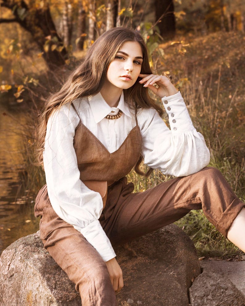 Woman wearing a white romantic linen blouse with puff sleeves, button cuffs, and vintage collar, styled with brown overalls in a fall forest setting. Victorian-inspired button-up shirt perfect for cottagecore outfits, feminine vintage looks, or countryside style. Ideal for photoshoots, layering, or holiday fashion. Ships fast from Brooklyn, USA — ready to ship in 24 hours with quick delivery.