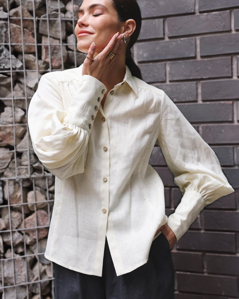 Woman wearing a tailored white linen blouse with puff sleeves, button cuffs, and pointed collar, styled with dark trousers in an urban setting. Feminine Victorian-inspired shirt with a clean, structured fit perfect for minimalist wardrobes, vintage office wear, or elegant everyday looks. Made from natural linen. Ships fast from Brooklyn, USA — ready to ship in 24 hours with quick delivery.