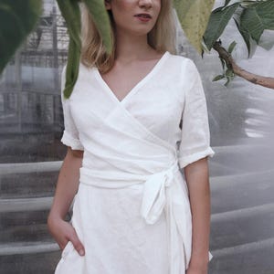 Ready to ship in 1 day from the USA, this handmade white linen wrap dress features a flattering V-neckline with delicate hand-finished topstitching, short sleeves with soft ruffle edging, and an adjustable waist tie. The breathable natural fabric and midi length make it perfect for a summer city hall wedding, rehearsal dinner, or as an elegant guest outfit. A minimalist, timeless piece designed for comfort and grace.