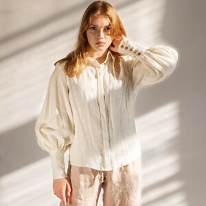 Model wearing a white linen blouse with puffed sleeves, buttoned cuffs, and soft Victorian collar, styled with beige linen trousers in warm natural light. Elegant cottagecore-inspired button-up shirt perfect for minimalist wardrobes, feminine vintage looks, or romantic office wear. Handmade and ready to ship in 24 hours from USA with fast Brooklyn-based shipping.