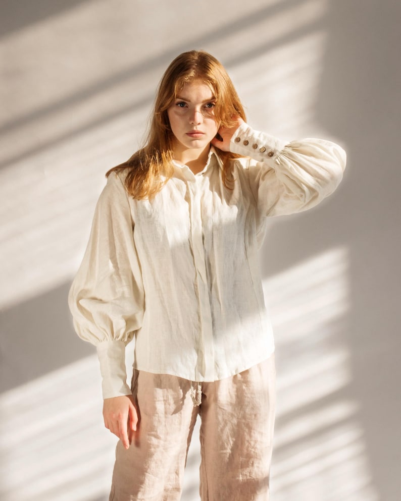 Model wearing a white linen blouse with puffed sleeves, buttoned cuffs, and soft Victorian collar, styled with beige linen trousers in warm natural light. Elegant cottagecore-inspired button-up shirt perfect for minimalist wardrobes, feminine vintage looks, or romantic office wear. Handmade and ready to ship in 24 hours from USA with fast Brooklyn-based shipping.