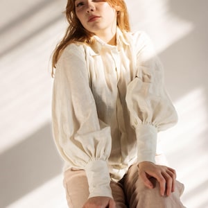 Woman wearing a romantic white linen blouse with voluminous puff sleeves, buttoned cuffs, and soft pleats, styled with beige linen trousers. Feminine and vintage-inspired, perfect for cottagecore looks, Victorian outfits, or slow fashion wardrobes. Ideal for elegant everyday wear or creative styling. Ships fast from Brooklyn, USA — ready to ship in 24 hours with quick delivery.