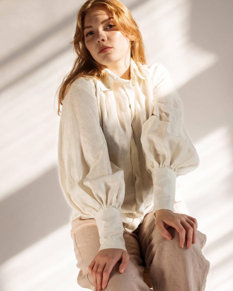 Woman wearing a romantic white linen blouse with voluminous puff sleeves, buttoned cuffs, and soft pleats, styled with beige linen trousers. Feminine and vintage-inspired, perfect for cottagecore looks, Victorian outfits, or slow fashion wardrobes. Ideal for elegant everyday wear or creative styling. Ships fast from Brooklyn, USA — ready to ship in 24 hours with quick delivery.