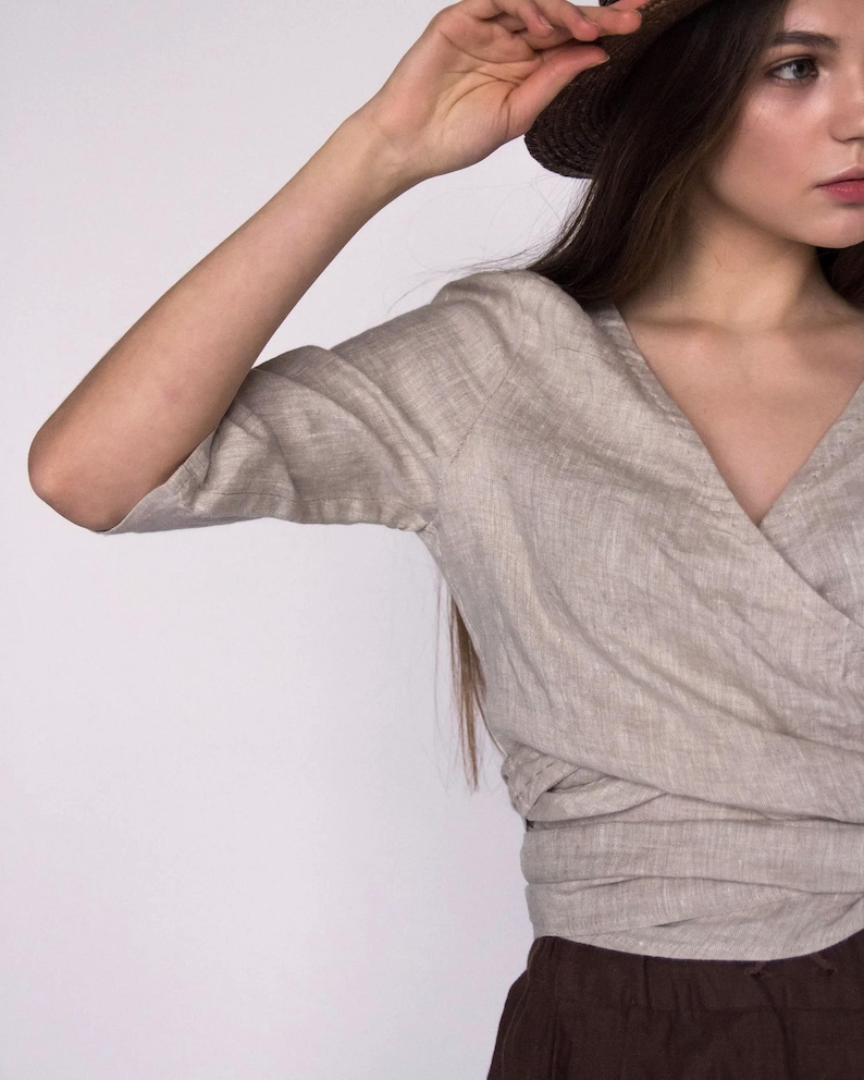 Close-up of a natural undyed linen wrap blouse showcasing the texture of the fabric, soft pleating, and visible hand-stitching detail along the neckline. Features a deep V-neck, relaxed fit, and rolled sleeves. Ships fast from the USA.