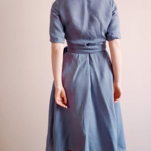 Back view of a woman wearing a dusty blue linen wrap dress with mid-length sleeves and a wide tie belt. The skirt falls just below the knees with a softly flared silhouette. The natural texture of linen and precise seams highlight the handcrafted quality of the garment. Ideal for everyday wear, garden gatherings, or vacation outfits. Ships from the USA within one business day.