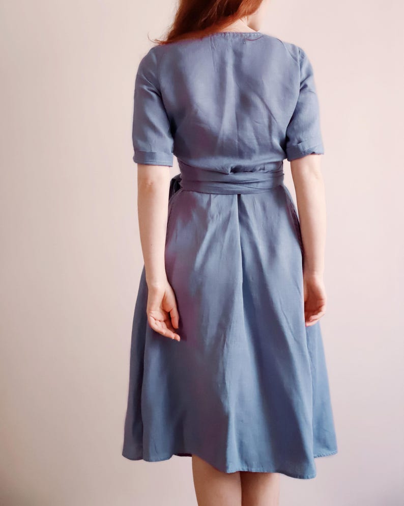 Back view of a woman wearing a dusty blue linen wrap dress with mid-length sleeves and a wide tie belt. The skirt falls just below the knees with a softly flared silhouette. The natural texture of linen and precise seams highlight the handcrafted quality of the garment. Ideal for everyday wear, garden gatherings, or vacation outfits. Ships from the USA within one business day.