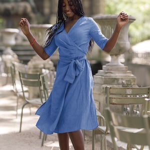 Woman joyfully twirling outdoors in a soft blue linen wrap dress with a V-neck, midi length, and adjustable waist tie. The dress moves gracefully as she spins, highlighting its lightweight fabric and feminine cut. A handmade decorative stitch along the neckline adds a special artisan touch. Perfect for summer walks, weekend getaways, or vacation photos. Ships quickly from the USA within one business day.