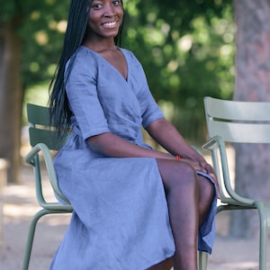 Woman sitting outdoors in a soft blue linen wrap dress with a tie waist and V-neckline. The dress flows gently around her legs, offering a feminine and elegant silhouette. A subtle handmade decorative stitch along the neckline adds a unique handcrafted touch. This comfortable and stylish dress is perfect for summer outings, vacations, and everyday wear. Ships from the USA within one business day.