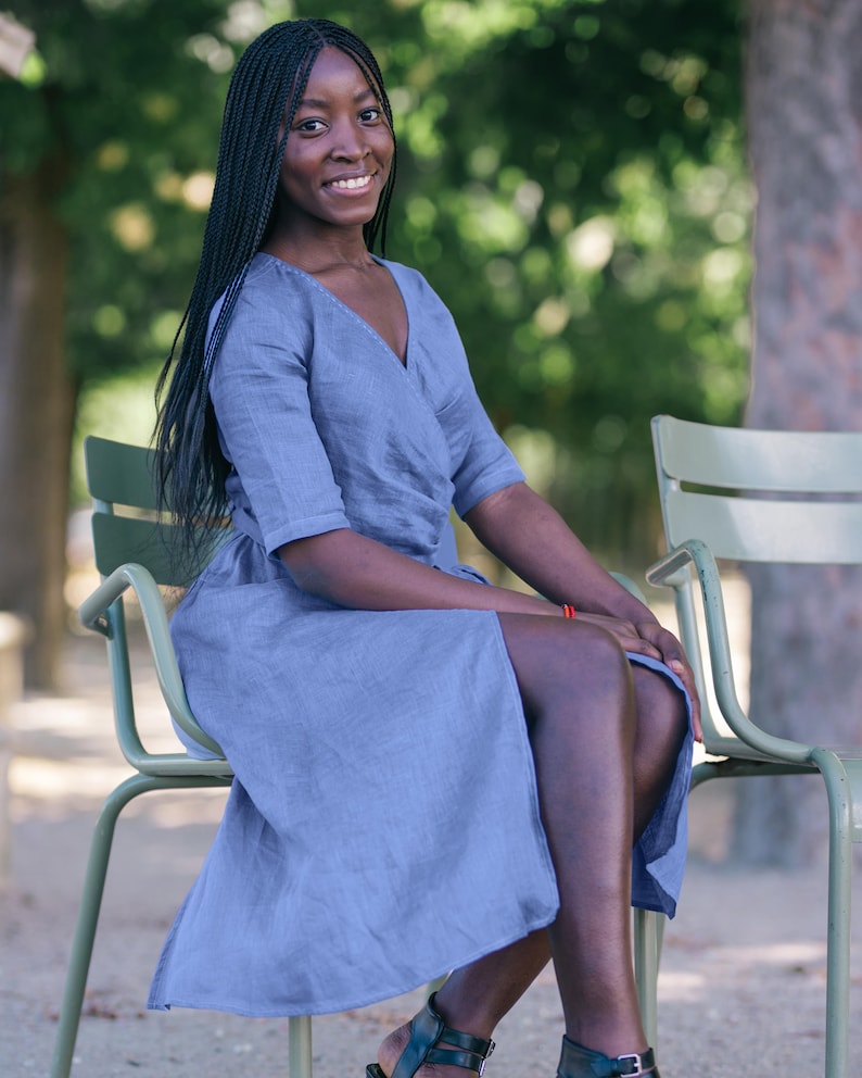 Woman sitting outdoors in a soft blue linen wrap dress with a tie waist and V-neckline. The dress flows gently around her legs, offering a feminine and elegant silhouette. A subtle handmade decorative stitch along the neckline adds a unique handcrafted touch. This comfortable and stylish dress is perfect for summer outings, vacations, and everyday wear. Ships from the USA within one business day.