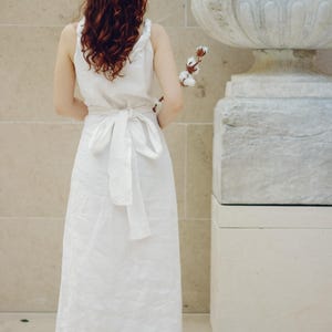 Woman with long wavy red hair wearing a white linen sleeveless wrap dress, tied at the back with a wide sash bow. Standing in front of a textured stone wall and classical sculpture, holding a cotton stem. The elegant midi-length dress is made of breathable natural linen—ideal for summer occasions, minimalist weddings, or garden events. Ships fast from the USA within 1 business day.