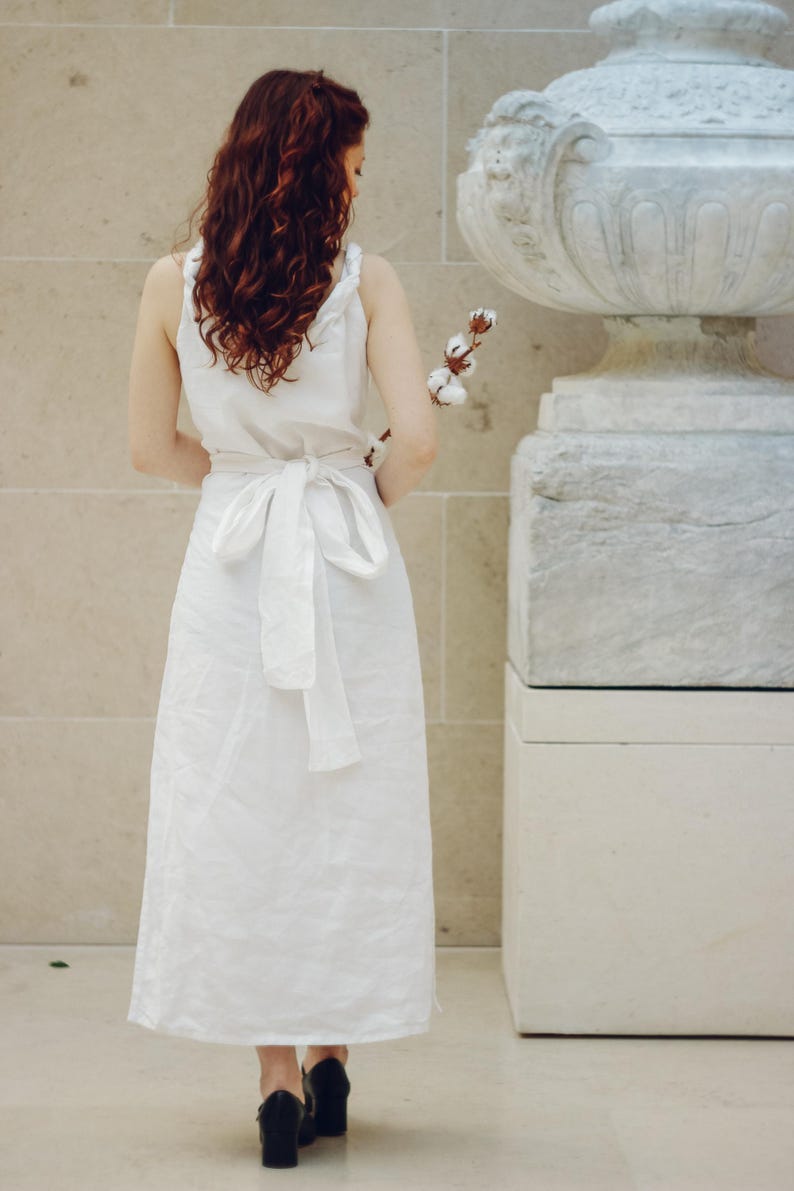 Woman with long wavy red hair wearing a white linen sleeveless wrap dress, tied at the back with a wide sash bow. Standing in front of a textured stone wall and classical sculpture, holding a cotton stem. The elegant midi-length dress is made of breathable natural linen—ideal for summer occasions, minimalist weddings, or garden events. Ships fast from the USA within 1 business day.