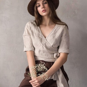 Linen wrap blouse for women made from softened natural linen, featuring a V-neckline with delicate hand stitching along the edge, a tie waist, and minimalist silhouette. Ideal for a creative summer wardrobe, city travel, or vacation styling. Ships quickly from the USA, dispatched within 1 day.