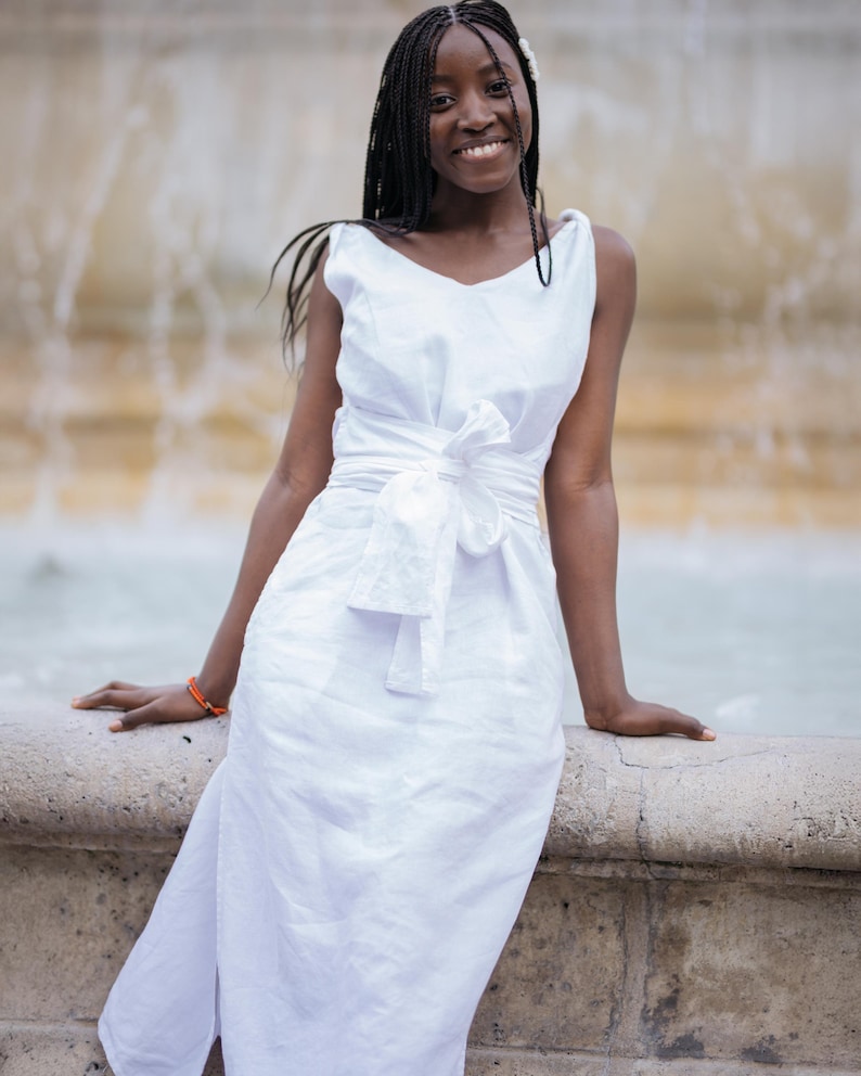 Woman wearing a white linen midi dress with wide V-neckline and twisted straps, standing by a fountain. The dress features a sewn-in waist tie, flattering fit, high side slits, and hidden pockets. Made from natural breathable linen, perfect for summer vacations, weddings, or beach dinners. Romantic and elegant look for women who value comfort and style. Ships fast from the USA within 1 business day.