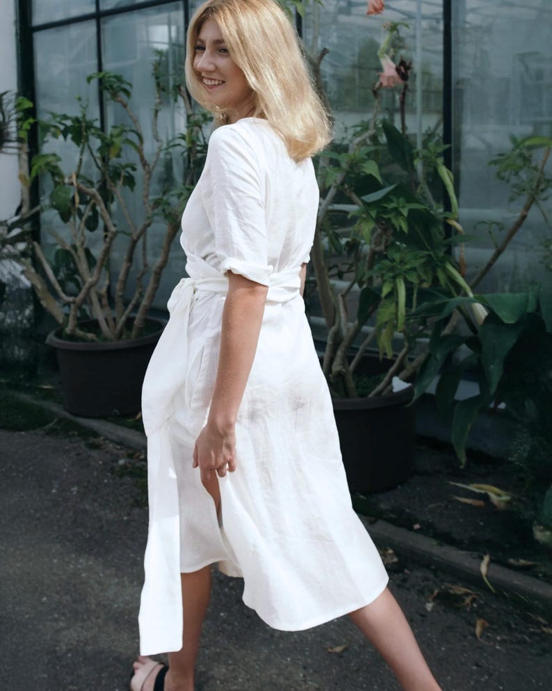 Ready to ship in 1 day from the USA, this handmade white linen wrap dress features a feminine silhouette with short sleeves, a flattering adjustable waist tie, and side slits for ease of movement and an airy summer feel. The midi length and breathable linen make it ideal for warm-weather events, city hall weddings, bridal showers, or effortless vacation outfits. Finished with soft hand-stitched details along the neckline.