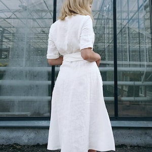 Back view of our handmade white linen wrap dress, ready to ship in 1 day from the USA. This breathable midi-length dress features a gently flared skirt, soft structured fit, and an adjustable tie waist for a flattering silhouette. Finished with hand-stitched details and short cuffed sleeves, it’s perfect for city hall weddings, summer gatherings, or effortless everyday elegance.