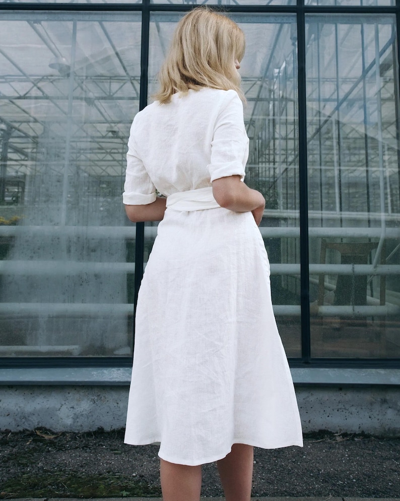 Back view of our handmade white linen wrap dress, ready to ship in 1 day from the USA. This breathable midi-length dress features a gently flared skirt, soft structured fit, and an adjustable tie waist for a flattering silhouette. Finished with hand-stitched details and short cuffed sleeves, it’s perfect for city hall weddings, summer gatherings, or effortless everyday elegance.