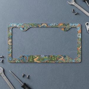 May include: A rectangular license plate frame featuring a colorful, repeating pattern of stylized mountains, evergreen trees, winding paths, and compass motifs in shades of blue, orange, and green against a dark gray background.