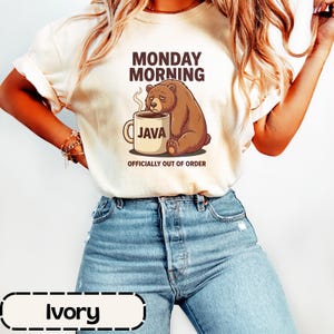 May include: An ivory t-shirt featuring a graphic of a tired brown bear leaning against a coffee mug labelled JAVA. The text above reads MONDAY MORNING and the text below reads OFFICIALLY OUT OF ORDER. The shirt is paired with blue denim jeans.
