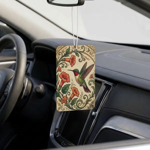 May include: A rectangular car air freshener hanging from a rearview mirror, featuring a detailed illustration of a hummingbird in flight surrounded by orange trumpet-shaped flowers and green leafy vines on a cream-colored background.