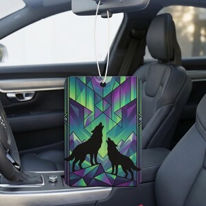 Puede incluir: Un ambientador de coche rectangular colgado del espejo retrovisor, con un diseño geométrico estilo vidriera de dos siluetas de lobos negros sobre un fondo vibrante verde, morado y azul con formas de montañas.