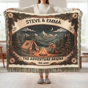 May include: A woven throw blanket featuring a camping scene with a tent, campfire, and two people sitting on a log under a starry sky. The text reads STEVE & EMMA, THE ADVENTURE BEGINS, and Est. 2026. The border includes pine cones and compasses.