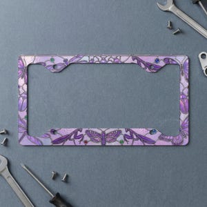 Puede incluir: Un marco de matrícula rectangular con un diseño de estilo vidriera en tonos morados y lavanda. El marco está decorado con ilustraciones de varios insectos, incluyendo polillas y mantis religiosas, acentuado por pequeños puntos coloridos.