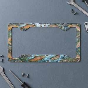 May include: A rectangular license plate frame featuring a colorful illustration of multiple trout swimming in a river with rocks and aquatic plants. The design depicts fish in shades of brown, orange, and green, set against a blue water background.