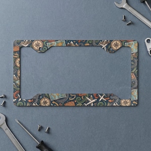 May include: A rectangular license plate frame featuring a repeating travel-themed pattern. The design includes illustrations of compasses, world maps, airplanes, mountains, and suitcases in shades of blue, orange, and tan on a dark background.