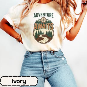 May include: An ivory t-shirt featuring a graphic print with the text ADVENTURE AWAITS. The design includes a compass, mountain peaks, a forest of pine trees, and a winding road contained within a circular border in shades of green and brown.