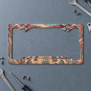 May include: A rectangular license plate frame featuring a desert landscape illustration with cacti, mesas, and a sun. The design incorporates geometric patterns in shades of orange, teal, and tan, with four mounting holes at the corners.