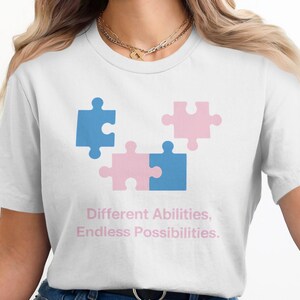 May include: White t-shirt featuring a graphic of four puzzle pieces in blue and pink. The text "Different Abilities, Endless Possibilities" is printed below the puzzle pieces. A casual, everyday tee.