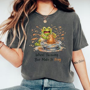 Puede incluir: Camiseta gris oscuro con un dibujo de una rana en un torno de alfarero. El texto dice "Wheel Throwing... But Make It Frog". La rana es verde y lleva un delantal blanco. La camiseta es de corte cómodo.