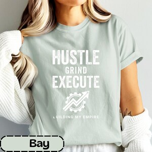 May include: A light sage green short-sleeve t-shirt featuring white text that reads HUSTLE GRIND EXECUTE above a graphic of a gear with an upward-pointing arrow and the text BUILDING MY EMPIRE at the bottom of the graphic design.