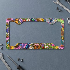 May include: A rectangular license plate frame featuring a vibrant, colorful pattern of various insects including butterflies, ladybugs, beetles, and dragonflies interspersed with white daisies and purple flowers against a pink background.