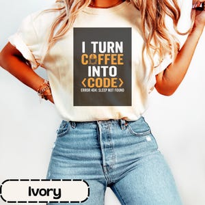 以下が含まれることがあります： アイボリーの半袖Tシャツ。中央にはダークグレーの長方形のグラフィックがあり、白とオレンジの文字で「I TURN COFFEE INTO <CODE>」および「ERROR 404: SLEEP NOT FOUND」と記されています。ライトブルーのデニムジーンズと組み合わされています。