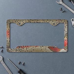 May include: A rectangular license plate frame featuring a detailed trout fish skin pattern. The design displays a textured surface with small black spots, golden-brown scales, and a prominent reddish-orange fin visible on the right side.