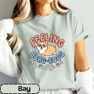 May include: A light sage green t-shirt featuring a graphic of a tan corgi wearing heart-shaped sunglasses. The text reads "FEELING CORG-EOUS" in pink and blue letters, accompanied by small colorful illustrations of dog bones and paw prints.