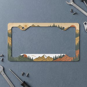 May include: A rectangular license plate frame featuring a printed landscape illustration. The design displays layered mountains in shades of tan, orange, and green, with dark green pine trees and a sun graphic against a light tan sky background.