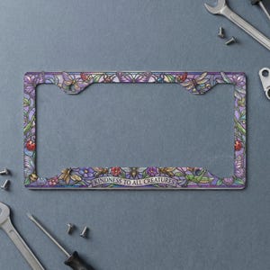 May include: A rectangular license plate frame featuring a stained glass style design with colorful butterflies, bees, ladybugs, and flowers. The bottom center of the frame displays the text "KINDNESS TO ALL CREATURES" in a clear, dark font.