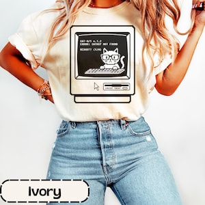 May include: An ivory t-shirt featuring a black graphic of a vintage computer monitor. The screen displays a pixelated cat wearing glasses at a keyboard with the text CAT-O/S v.1.2, ERROR: CATNIP NOT FOUND, REBOOT? (Y/N), and INSERT TREAT.