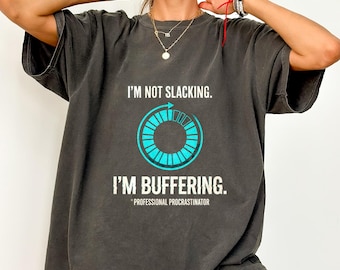 May include: A charcoal gray t-shirt featuring a graphic of a cyan circular loading icon. The text reads: I'M NOT SLACKING. I'M BUFFERING. PROFESSIONAL PROCRASTINATOR. The shirt has a crew neckline and short sleeves, with a relaxed, loose-fitting silhouette.