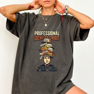Pode incluir: Uma t-shirt cinzento-carvão com um gráfico de uma pessoa com uma pilha de vários chapéus na cabeça. O texto diz PROFESSIONAL IDENTITY CRISIS e WHICH HAT AM I WEARING TODAY? Os chapéus estão rotulados com cargos como Project Manager.